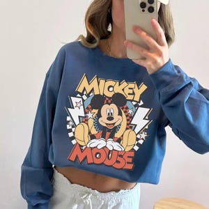 May include: A cropped, blue sweatshirt featuring a graphic of Mickey Mouse. The design includes the words "Mickey Mouse" in yellow and red, with lightning bolt and star accents. The cartoon character is depicted in a seated position.