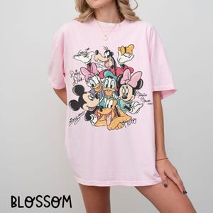 May include: A light pink t-shirt featuring a colorful graphic of Disney characters, including Mickey Mouse, Minnie Mouse, Donald Duck, and Goofy. The shirt has short sleeves and a crew neck. The word "BLOSSOM" is printed at the bottom.