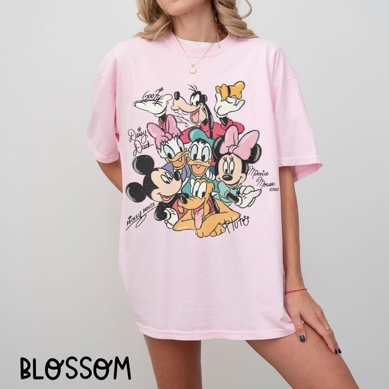 May include: A light pink t-shirt featuring a colorful graphic of Disney characters, including Mickey Mouse, Minnie Mouse, Donald Duck, and Goofy. The shirt has short sleeves and a crew neck. The word "BLOSSOM" is printed at the bottom.