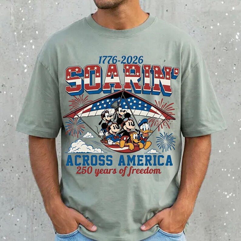 K&ouml;nnte beinhalten: Ein salbeigr&uuml;nes T-Shirt mit einer Grafik von Mickey Maus, Donald Duck, Goofy und Pluto auf einem H&auml;ngegleiter. Der Text lautet 1776-2026 SOARIN' ACROSS AMERICA 250 years of freedom, erg&auml;nzt durch ein rot-wei&szlig;-blaues Flaggenmuster und Feuerwerk.