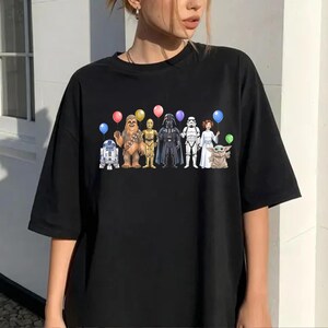 May include: Black t-shirt with a vibrant print of Star Wars characters, each holding a balloon. The design includes R2-D2, Chewbacca, C-3PO, Darth Vader, a Stormtrooper, Princess Leia, and Grogu.