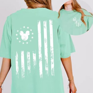 May include: A light green t-shirt featuring a distressed white American flag design. The flag incorporates a Mickey Mouse head in place of the stars. The shirt has short sleeves and a relaxed fit, suitable for casual wear.