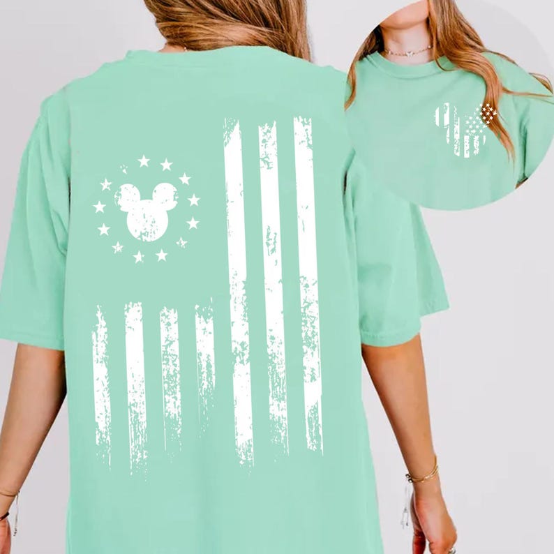 May include: A light green t-shirt featuring a distressed white American flag design. The flag incorporates a Mickey Mouse head in place of the stars. The shirt has short sleeves and a relaxed fit, suitable for casual wear.