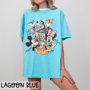 May include: Lagoon blue t-shirt featuring a graphic of Disney characters including Mickey Mouse, Minnie Mouse, Donald Duck, Daisy Duck, Goofy, and Pluto. The shirt has short sleeves and a crew neck. The text "Mickey Mouse" and "Minnie Mouse" are written on the shirt.