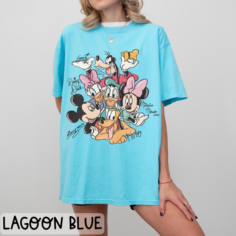 May include: Lagoon blue t-shirt featuring a graphic of Disney characters including Mickey Mouse, Minnie Mouse, Donald Duck, Daisy Duck, Goofy, and Pluto. The shirt has short sleeves and a crew neck. The text "Mickey Mouse" and "Minnie Mouse" are written on the shirt.
