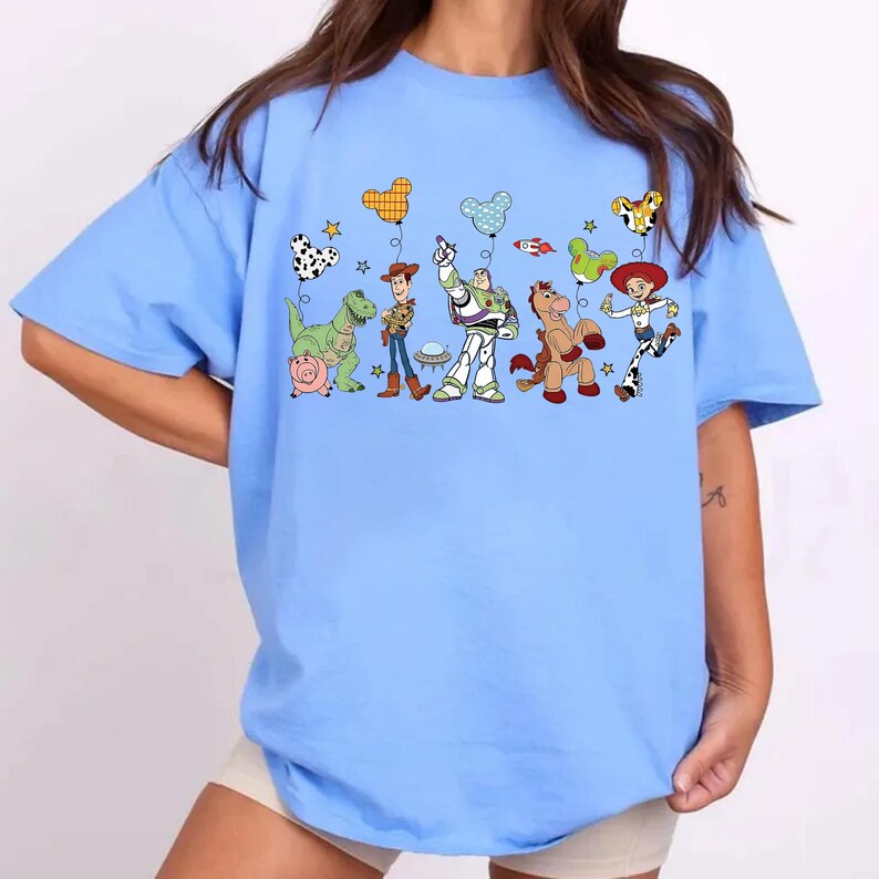 May include: Light blue t-shirt featuring a colorful graphic of animated characters and balloons. The design includes a dinosaur, a pig, a cowboy, a space ranger, and a horse. The balloons have various patterns and shapes, adding to the playful theme.