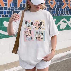 May include: White t-shirt featuring a colorful graphic of Minnie Mouse in four different poses. The graphic has a pastel color scheme with light blue, pink, and yellow squares. The shirt is a casual, oversized fit.