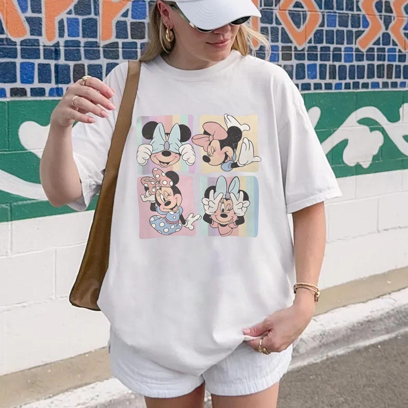 May include: White t-shirt featuring a colorful graphic of Minnie Mouse in four different poses. The graphic has a pastel color scheme with light blue, pink, and yellow squares. The shirt is a casual, oversized fit.