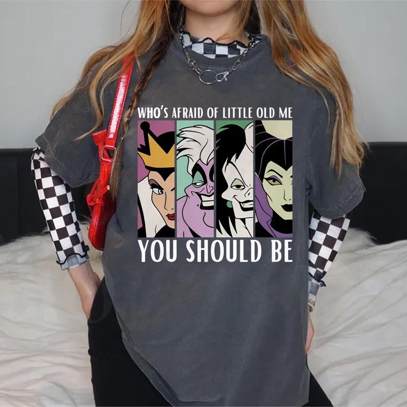 Puede incluir: Camiseta gris oscuro con un gr&aacute;fico de cuatro villanos de dibujos animados y el texto "WHO'S AFRAID OF LITTLE OLD ME YOU SHOULD BE". La camiseta se usa sobre una camisa de manga larga a cuadros en blanco y negro.