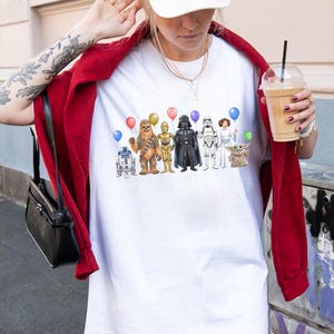 May include: White t-shirt with a colourful illustration of Star Wars characters holding balloons. The characters include R2-D2, Chewbacca, C-3PO, Darth Vader, a Stormtrooper, Princess Leia, and Grogu.