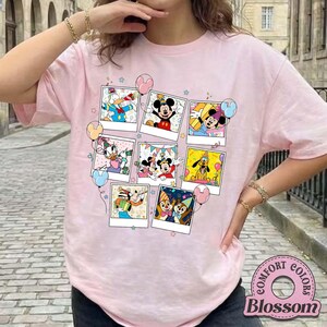 Puede incluir: Camiseta rosa claro con un collage de im&aacute;genes de personajes de dibujos animados en marcos polaroid. Los personajes incluyen Mickey Mouse, Minnie Mouse, Donald Duck y Goofy. La camiseta tiene cuello redondo y mangas cortas. El logo de la marca "Comfort Colors Blossom" es visible.