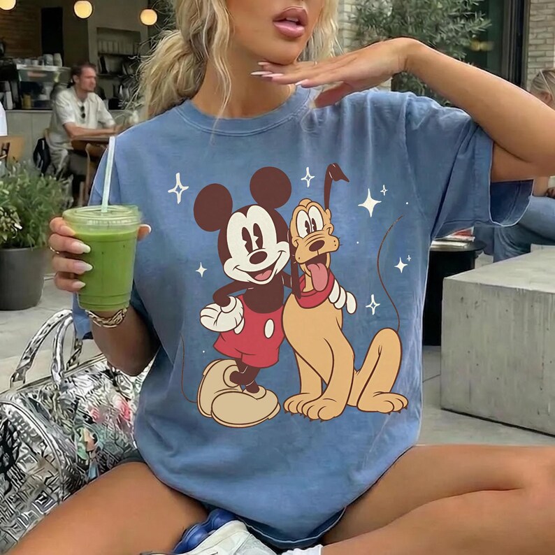 May include: A light blue t-shirt with a graphic of Mickey Mouse and Pluto. Mickey is in red shorts, and Pluto has a red collar. The shirt has a vintage wash and a crew neck.