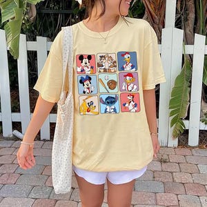May include: A light yellow t-shirt featuring a grid of cartoon characters in baseball-themed poses. The characters include Mickey Mouse, Chip and Dale, Donald Duck, Minnie Mouse, a baseball bat and glove, and Stitch.
