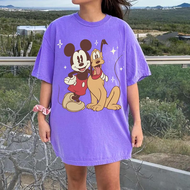 May include: A lilac t-shirt with a graphic of Mickey Mouse and Pluto. Mickey is in red shorts and Pluto has a red collar. The design includes star accents. The shirt is a casual, oversized style.