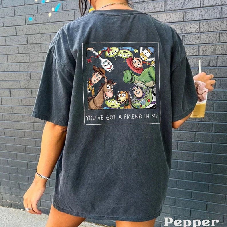 May include: A dark gray t-shirt featuring a colorful graphic of characters from the animated film "Toy Story." The graphic is framed with the text "YOU'VE GOT A FRIEND IN ME." The shirt has short sleeves and a relaxed fit.