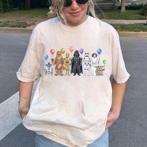 May include: Cream-coloured t-shirt with a vibrant illustration of Star Wars characters, each holding a balloon. The characters depicted are R2-D2, Chewbacca, C-3PO, Darth Vader, a Stormtrooper, Princess Leia, and Grogu.