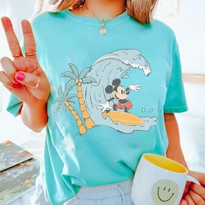 May include: Turquoise t-shirt featuring a cartoon Mickey Mouse surfing a wave. The design includes palm trees and a yellow surfboard. The shirt is worn by a person holding a mug with a smiley face.