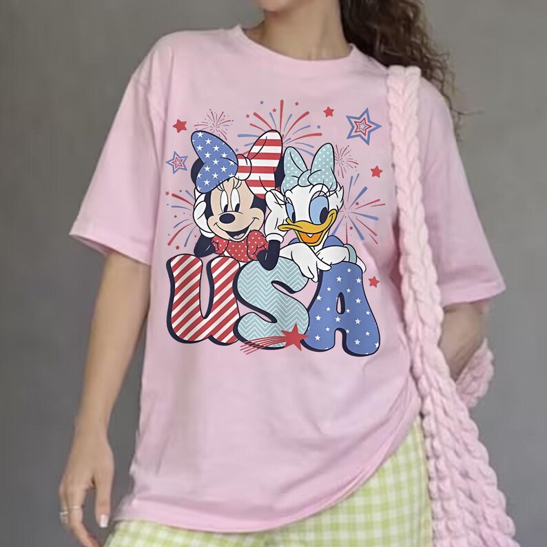 May include: Pink t-shirt featuring Minnie Mouse and Daisy Duck with the word "USA" in red, white, and blue. The graphic includes fireworks and stars. The shirt has short sleeves and a crew neck.
