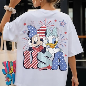 May include: White t-shirt featuring Minnie Mouse and Daisy Duck with a patriotic "USA" design. The graphic includes red, white, and blue elements, fireworks, and stars. The shirt is a casual, oversized style.
