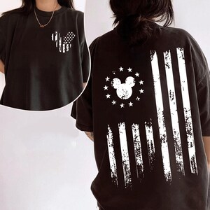 May include: Black t-shirt with a distressed white graphic of a Mickey Mouse-shaped American flag on the front and back. The back design features a Mickey Mouse head surrounded by stars and stripes.