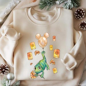 May include: Cream-colored sweatshirt featuring a cartoon chameleon holding a flower and a Mickey Mouse-shaped balloon. The design includes floating lanterns and flowers in shades of orange and yellow. The sweatshirt has long sleeves and a crew neck.