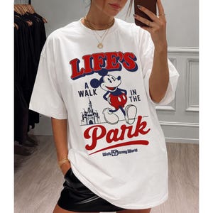 Puede incluir: Camiseta blanca con un dise&ntilde;o de Mickey Mouse y el texto "LIFE'S A WALK IN THE PARK" en rojo y azul. El dise&ntilde;o incluye un castillo y las palabras "Walt Disney World".
