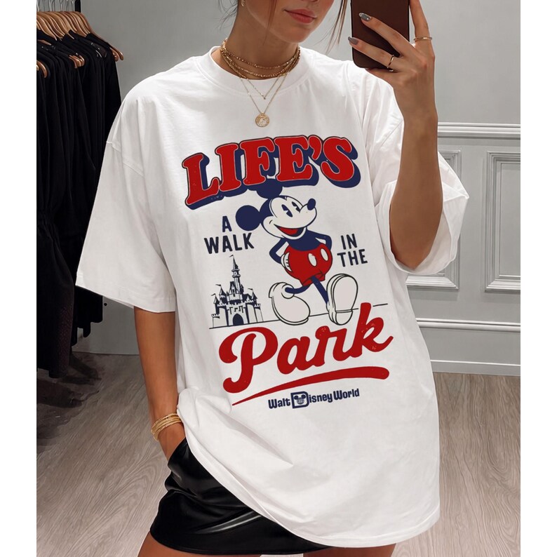 Puede incluir: Camiseta blanca con un dise&ntilde;o de Mickey Mouse y el texto "LIFE'S A WALK IN THE PARK" en rojo y azul. El dise&ntilde;o incluye un castillo y las palabras "Walt Disney World".