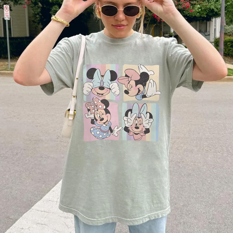 May include: Sage green t-shirt featuring four squares with different Minnie Mouse illustrations. Each square has a pastel background. The shirt is a relaxed fit and has short sleeves.