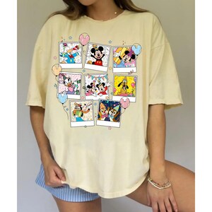 Puede incluir: Una camiseta amarillo claro con un collage de im&aacute;genes de personajes de dibujos animados en marcos estilo polaroid. Las im&aacute;genes incluyen Mickey Mouse, Minnie Mouse, Donald Duck y Pluto, con globos y estrellas de colores. La camiseta tiene mangas cortas y un corte holgado.