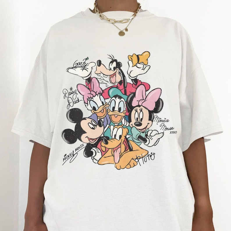 May include: White t-shirt featuring a colorful graphic of Disney characters including Mickey Mouse, Minnie Mouse, Donald Duck, Daisy Duck, Goofy, and Pluto. The shirt has short sleeves and a crew neck. The characters' names are written in cursive.