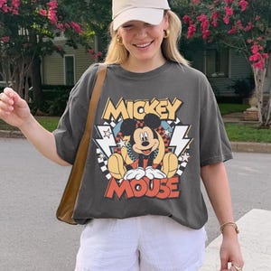 May include: Dark gray t-shirt featuring a graphic of Mickey Mouse. The design includes the words "Mickey Mouse" in a retro font, with lightning bolts and stars. The shirt is worn with white shorts and a tan shoulder bag.
