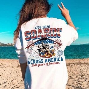 K&ouml;nnte beinhalten: Ein wei&szlig;es Langarmshirt mit einer Grafik von Zeichentrickfiguren in einem H&auml;ngegleiter mit US-Flaggenmuster. Der Text lautet 1776-2026 SOARIN' ACROSS AMERICA 250 years of freedom, erg&auml;nzt durch rote und blaue Feuerwerksdarstellungen.