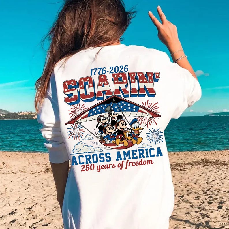 K&ouml;nnte beinhalten: Ein wei&szlig;es Langarmshirt mit einer Grafik von Zeichentrickfiguren in einem H&auml;ngegleiter mit US-Flaggenmuster. Der Text lautet 1776-2026 SOARIN' ACROSS AMERICA 250 years of freedom, erg&auml;nzt durch rote und blaue Feuerwerksdarstellungen.