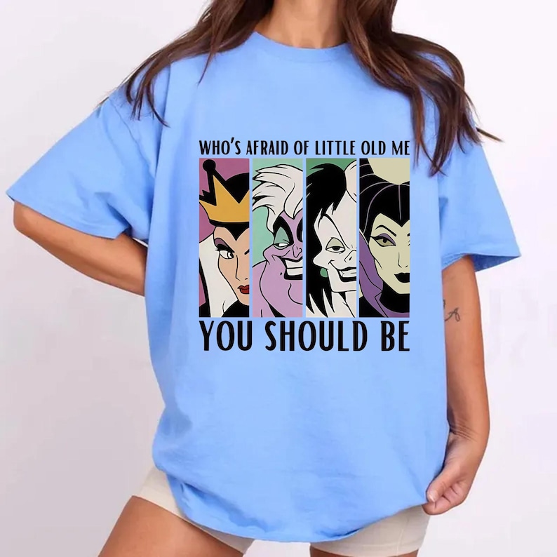 Puede incluir: Camiseta azul claro con un estampado de cuatro villanas de dibujos animados: la Reina Malvada, &Uacute;rsula, Cruella de Vil y Mal&eacute;fica. El texto dice: "WHO'S AFRAID OF LITTLE OLD ME YOU SHOULD BE".
