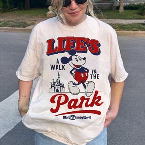 Puede incluir: Camiseta color crema con un gr&aacute;fico de Mickey Mouse caminando frente a un castillo. El texto dice "Life's a Walk in the Park" en rojo y azul, con "Walt Disney World" debajo. La camiseta tiene mangas cortas.