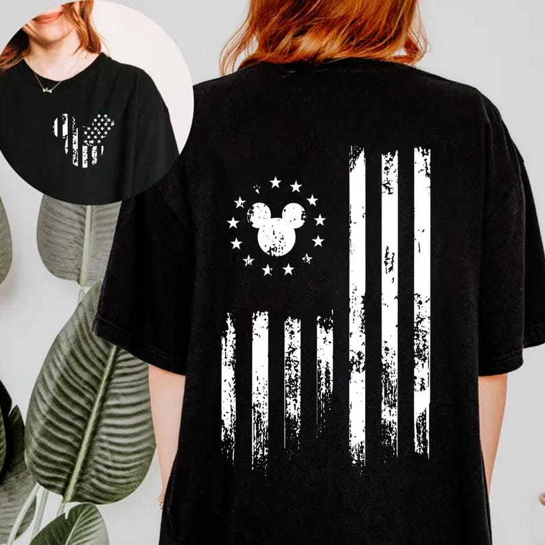 May include: Black t-shirt featuring a distressed white graphic of the American flag with a Mickey Mouse head in place of the stars. The design is on the front and back of the shirt. The shirt is a casual style.