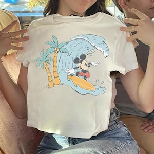 May include: Off-white t-shirt featuring a cartoon illustration of Mickey Mouse surfing a wave. The design includes palm trees and a surfboard. The shirt has short sleeves and a crew neck.