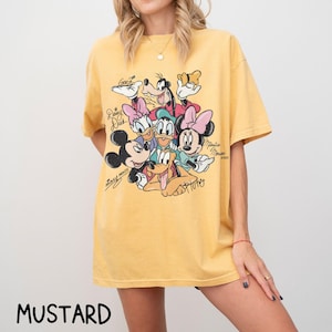 May include: Mustard-colored t-shirt featuring a graphic of Disney characters including Mickey Mouse, Minnie Mouse, Donald Duck, Daisy Duck, Goofy, and Pluto. The shirt has a crew neck and short sleeves. The word "MUSTARD" is printed at the bottom.