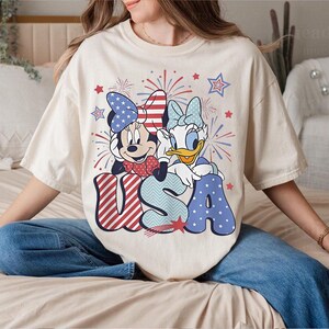 May include: Cream-colored t-shirt featuring Minnie Mouse and Daisy Duck with the word "USA" in red, white, and blue. The design includes fireworks and stars, celebrating a patriotic theme. The shirt is a relaxed fit.