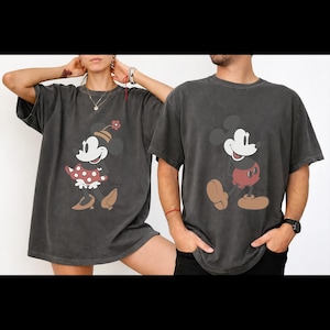 May include: Two charcoal gray t-shirts featuring Disney's Mickey and Minnie Mouse. Minnie is wearing a red polka dot dress and a flower hat. Mickey is wearing red shorts. Both characters are smiling.