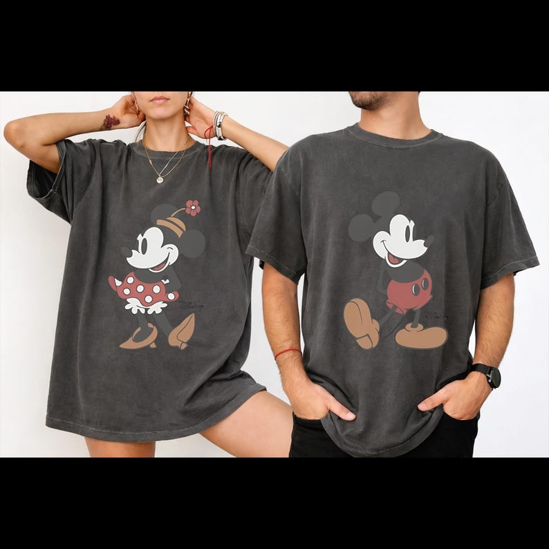 May include: Two charcoal gray t-shirts featuring Disney's Mickey and Minnie Mouse. Minnie is wearing a red polka dot dress and a flower hat. Mickey is wearing red shorts. Both characters are smiling.