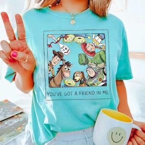 May include: A light blue t-shirt featuring a framed graphic of Toy Story characters, including Woody, Buzz Lightyear, and Rex. The text "YOU'VE GOT A FRIEND IN ME" is printed below the graphic. A white mug with a yellow interior and a smiley face is also visible.