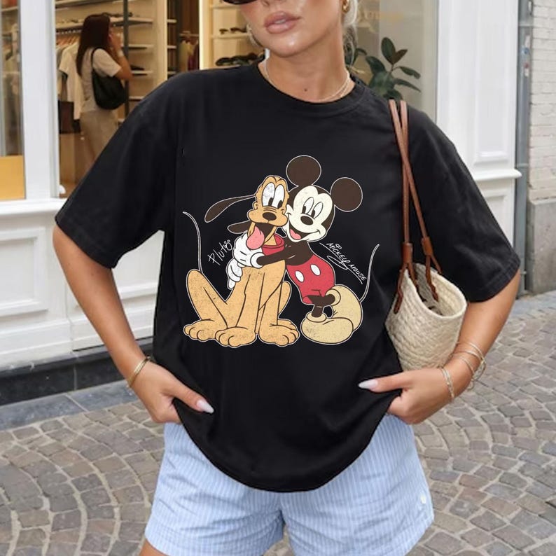 May include: Black t-shirt featuring a graphic of Mickey Mouse and Pluto embracing. Mickey Mouse is wearing red shorts and yellow shoes. Pluto is tan. The words "Pluto" and "Mickey Mouse" are written on the shirt.