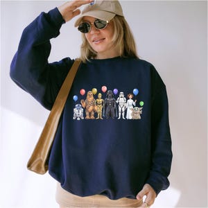 May include: Navy blue sweatshirt with a Star Wars character design. The design includes R2-D2, Chewbacca, C-3PO, Darth Vader, a Stormtrooper, Princess Leia, and Grogu, each holding a balloon. A casual, comfortable style.