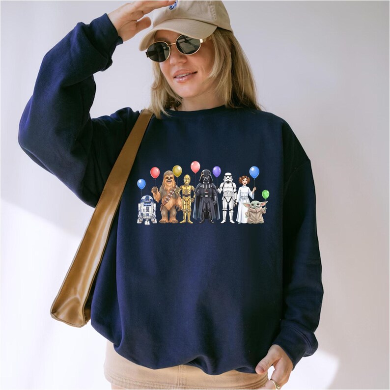 May include: Navy blue sweatshirt with a Star Wars character design. The design includes R2-D2, Chewbacca, C-3PO, Darth Vader, a Stormtrooper, Princess Leia, and Grogu, each holding a balloon. A casual, comfortable style.