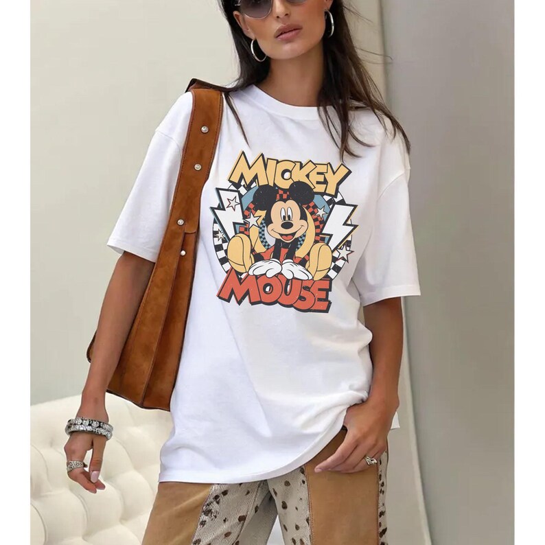May include: White t-shirt featuring a colorful graphic of Mickey Mouse with the text "Mickey Mouse". The design includes a checkered background and lightning bolt accents. The shirt has short sleeves.