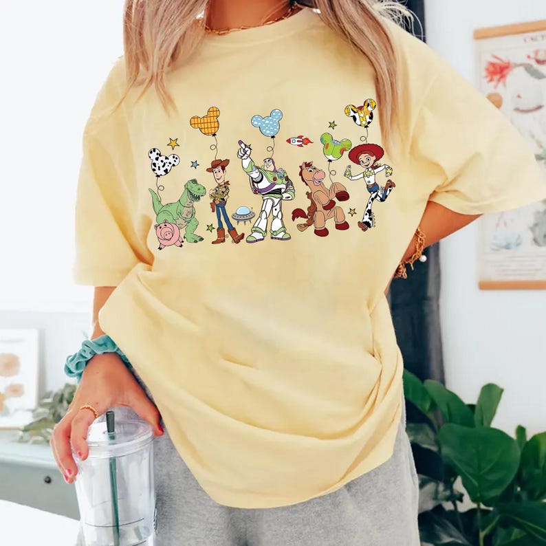 May include: A light yellow t-shirt featuring a colorful graphic of characters from a popular animated movie. The design includes a dinosaur, a cowboy, a space ranger, a horse, and a cowgirl, all connected by balloon-shaped accents.