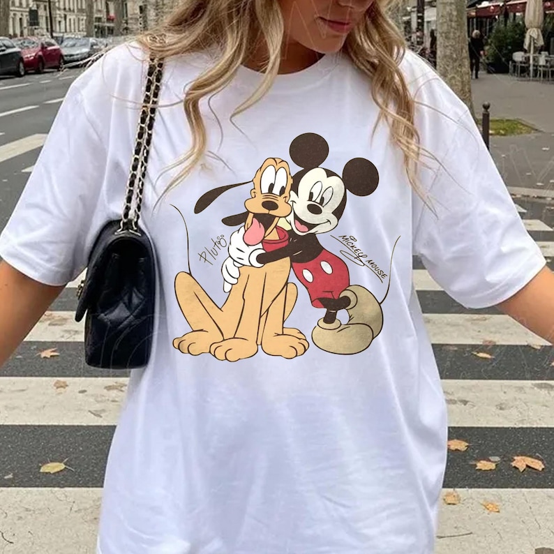 May include: White t-shirt featuring a cartoon graphic of Mickey Mouse hugging Pluto. Mickey Mouse is wearing red shorts and Pluto is tan. The words "Pluto" and "Mickey Mouse" are written on the shirt.