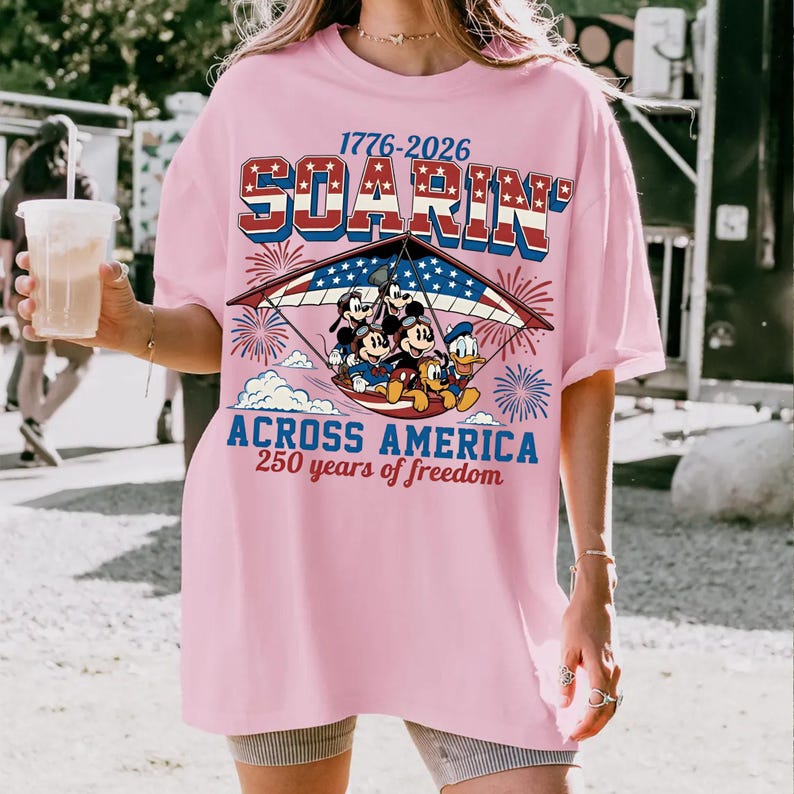 K&ouml;nnte beinhalten: Ein hellrosa T-Shirt mit einer Grafik von Zeichentrickfiguren beim Drachenfliegen im patriotischen US-Flaggen-Design. Der Text lautet 1776-2026 SOARIN' ACROSS AMERICA 250 years of freedom, umgeben von Feuerwerk und Sternen in Rot, Wei&szlig; und Blau.