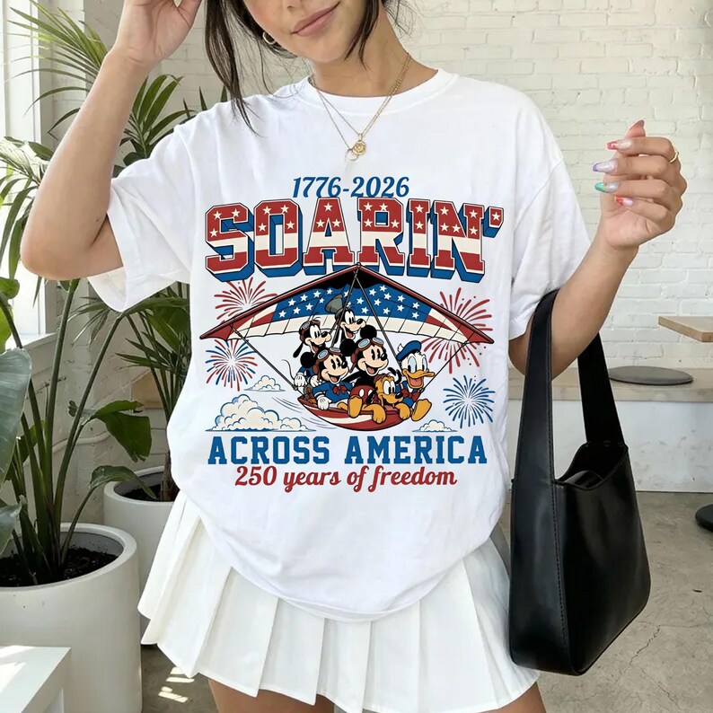 K&ouml;nnte beinhalten: Ein wei&szlig;es Kurzarm-T-Shirt mit einer Grafik von Mickey Maus, Donald Duck und Goofy in einem H&auml;ngegleiter. Der Text lautet 1776-2026 SOARIN' ACROSS AMERICA 250 years of freedom, erg&auml;nzt durch rot-wei&szlig;-blaue Sterne, Streifen und Feuerwerk.